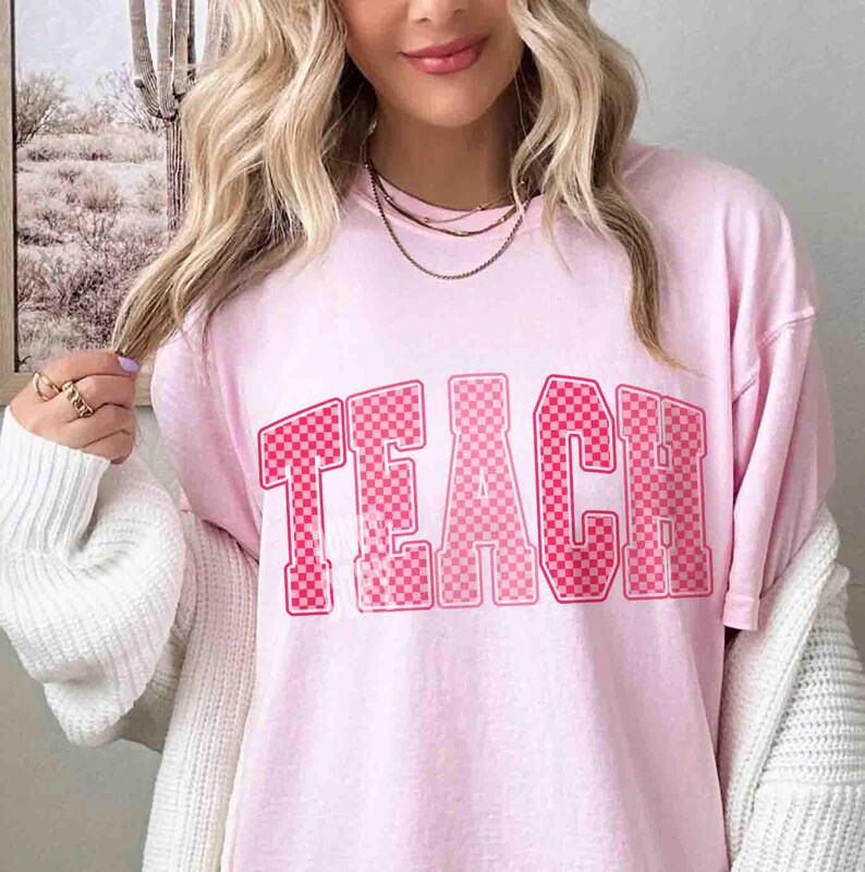 K&ouml;nnte beinhalten: Hellrosa T-Shirt mit dem Wort "TEACH" in einem rosa karierten Design. Das Shirt hat einen Rundhalsausschnitt und kurze &Auml;rmel. Der Stoff wirkt weich und bequem. Der Stil ist l&auml;ssig.