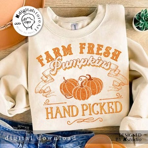 May include: Cream-colored sweatshirt with the text "FARM FRESH PUMPKINS HAND PICKED" in orange. The design includes three orange pumpkins and decorative banners. The sweatshirt is folded, with a pair of blue jeans visible.