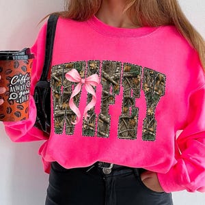 Puede incluir: Una sudadera rosa brillante con la palabra "Wifey" en un patr&oacute;n de camuflaje, acentuada con un lazo rosa. La sudadera es de un color vibrante y tiene mangas largas. Se ve una taza de caf&eacute; con el texto "Coffee Always a Good Idea".