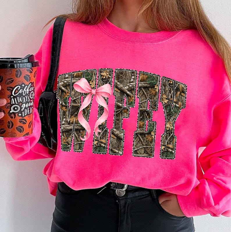 Puede incluir: Una sudadera rosa brillante con la palabra "Wifey" en un patr&oacute;n de camuflaje, acentuada con un lazo rosa. La sudadera es de un color vibrante y tiene mangas largas. Se ve una taza de caf&eacute; con el texto "Coffee Always a Good Idea".