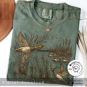 May include: Sage green t-shirt featuring a detailed illustration of ducks in a marsh setting. The design includes ducks in flight and swimming, cattails, and the text "digital download". A silver necklace with a pendant is draped across the shirt.