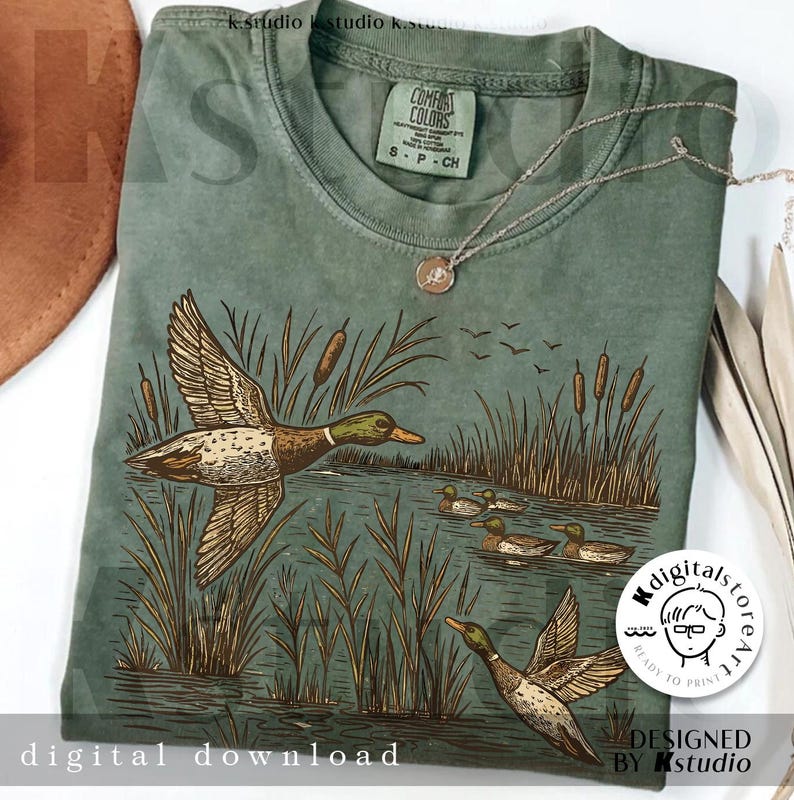 May include: Sage green t-shirt featuring a detailed illustration of ducks in a marsh setting. The design includes ducks in flight and swimming, cattails, and the text "digital download". A silver necklace with a pendant is draped across the shirt.