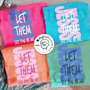 May include: Four colorful t-shirts, each displaying the text "LET THEM SEE YOU IN ME." The shirts are in pink, teal, orange, and green, with the text in contrasting colors. A small logo is visible in the center.