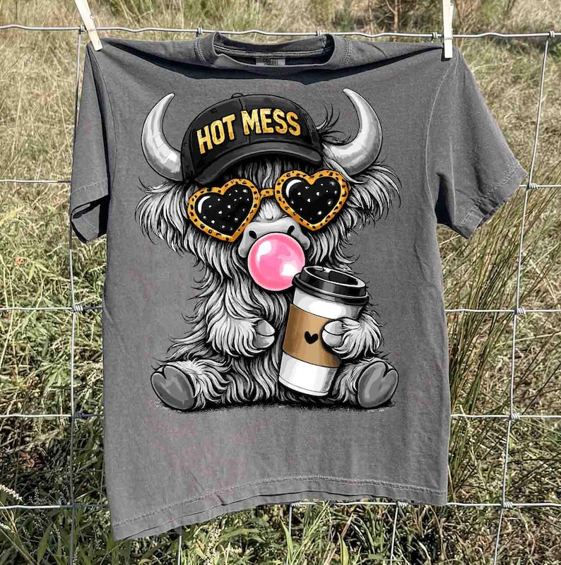 K&ouml;nnte beinhalten: Graues T-Shirt mit einem Cartoon-Highland-Rind, das eine schwarze Kappe mit dem Text "HOT MESS", herzf&ouml;rmige Sonnenbrille tr&auml;gt und eine Blase bl&auml;st. Das Rind h&auml;lt eine Kaffeetasse. Das T-Shirt h&auml;ngt an einer W&auml;scheleine.