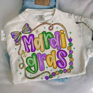 May include: Cream-colored t-shirt with a colorful "Mardi Gras" design. The design features the words "Mardi Gras" in purple, yellow, and green bubble letters, with a bow, fleur-de-lis, and bead accents. The shirt is folded on top of blue jeans.