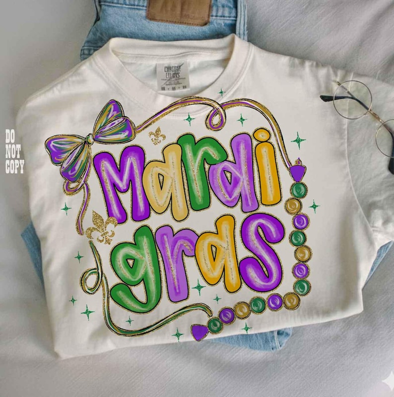 May include: Cream-colored t-shirt with a colorful "Mardi Gras" design. The design features the words "Mardi Gras" in purple, yellow, and green bubble letters, with a bow, fleur-de-lis, and bead accents. The shirt is folded on top of blue jeans.