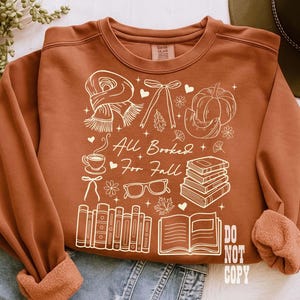 May include: Rust-colored sweatshirt with a white graphic design featuring a scarf, bow, pumpkin, books, coffee cup, glasses, and the text "All Booked For Fall." The design also includes the text "DO NOT COPY."
