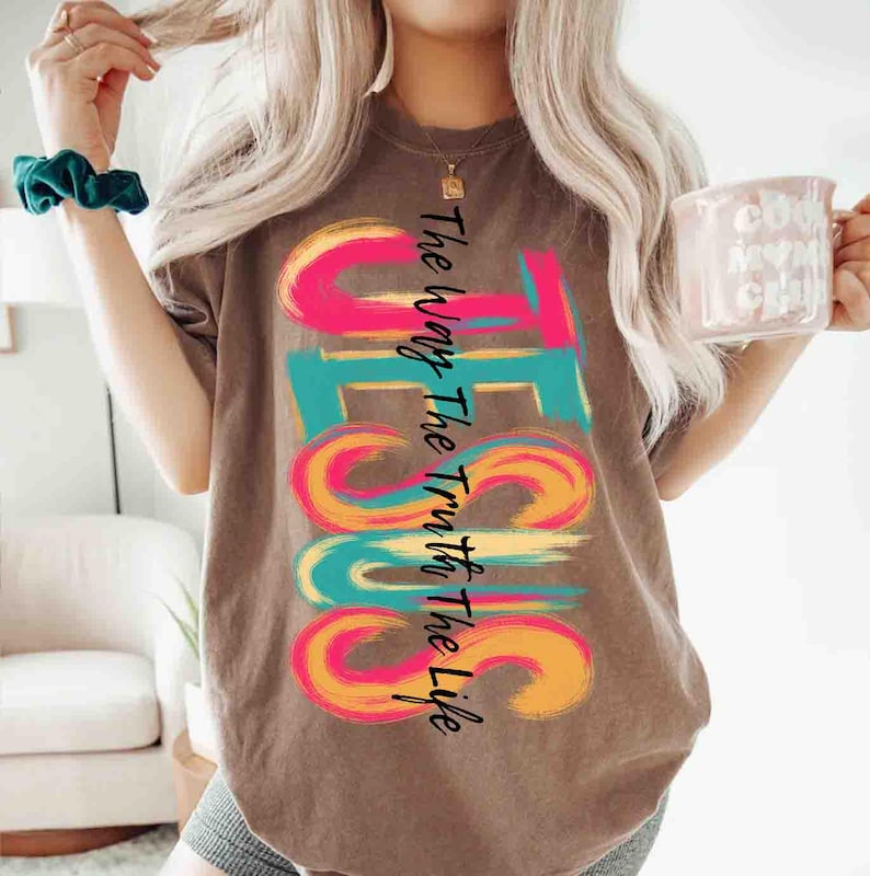 May include: Brown t-shirt with the word "JESUS" in large, colorful, brushstroke letters. The text "The Way The Truth The Life" is printed vertically along the side. The shirt is a casual style.