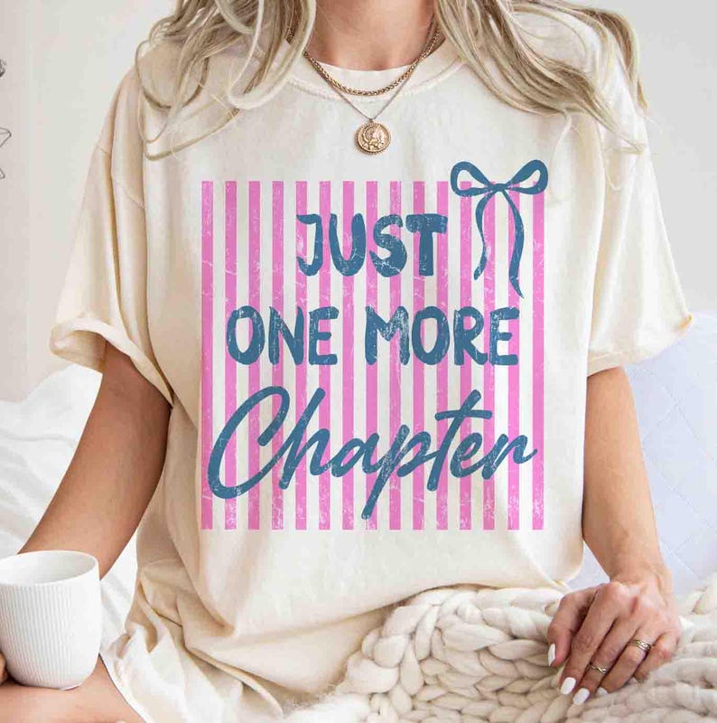 Puede incluir: Camiseta color crema con el texto azul "JUST ONE MORE CHAPTER" sobre un fondo de rayas rosas y blancas. Un lazo azul est&aacute; encima del texto. La camiseta es de estilo casual y c&oacute;modo.