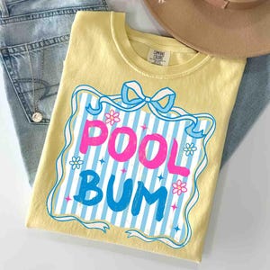 May include: A yellow t-shirt featuring the words "POOL BUM" in pink and blue, set within a blue and white striped design with flowers and a bow. The shirt is placed on blue jeans, with a tan hat visible.