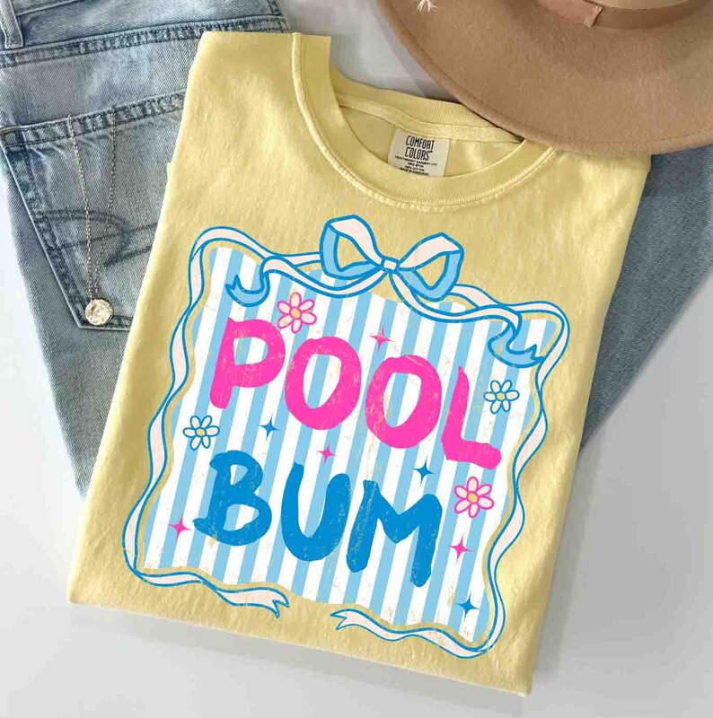 May include: A yellow t-shirt featuring the words "POOL BUM" in pink and blue, set within a blue and white striped design with flowers and a bow. The shirt is placed on blue jeans, with a tan hat visible.