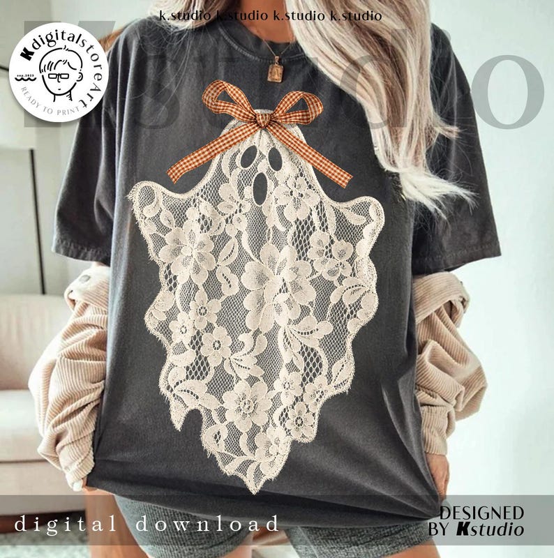 May include: A charcoal gray short-sleeve t-shirt featuring a large graphic of a ghost shape made from cream-colored floral lace. The ghost is topped with a brown and white checkered bow. The shirt has a crew neckline and a relaxed fit.