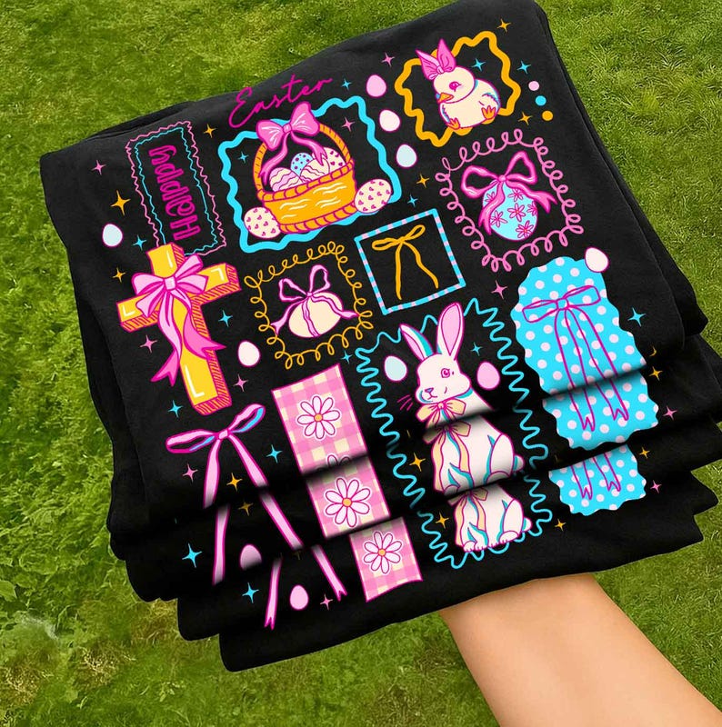 May include: A stack of black t-shirts featuring colorful Easter-themed designs. The designs include a cross, Easter eggs, bunnies, bows, and the word "Happy" and "Easter". The shirts are on a green grassy surface.
