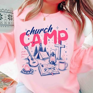 K&ouml;nnte beinhalten: Rosa Sweatshirt mit einem grafischen Design mit den Worten "church CAMP" in Blau und Pink. Das Design umfasst eine Gitarre, ein Zelt, ein Lagerfeuer, ein Kreuz, eine Laterne, ein offenes Buch und eine Kaffeetasse. Die Worte "faith friends fun" sind ebenfalls enthalten.