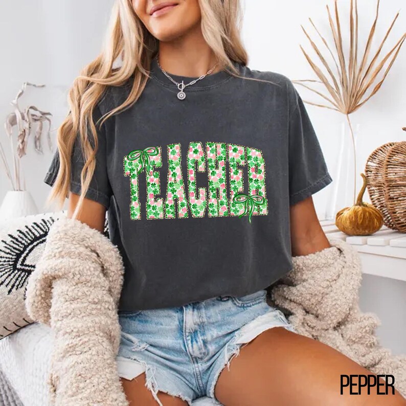 May include: Dark gray t-shirt with the word "TEACHER" in a green and pink shamrock pattern. The shirt has short sleeves and a crew neck. The design includes green bows. The model is wearing denim shorts and a cream-colored sweater.
