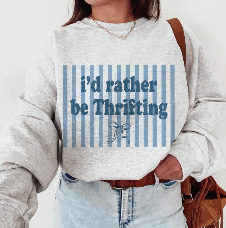 May include: A light gray sweatshirt with the text "I'd rather be Thrifting" in blue. The text is set against a blue and white striped background with a bow graphic. The sweatshirt has long sleeves and a crew neck.