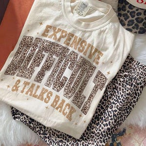 May include: Cream-coloured t-shirt with the text "EXPENSIVE, DIFFICULT, & TALKS BACK" in brown and leopard print. The shirt is paired with leopard print trousers. The Comfort Colors brand tag is visible. The outfit is laid out on a white fur-like surface.