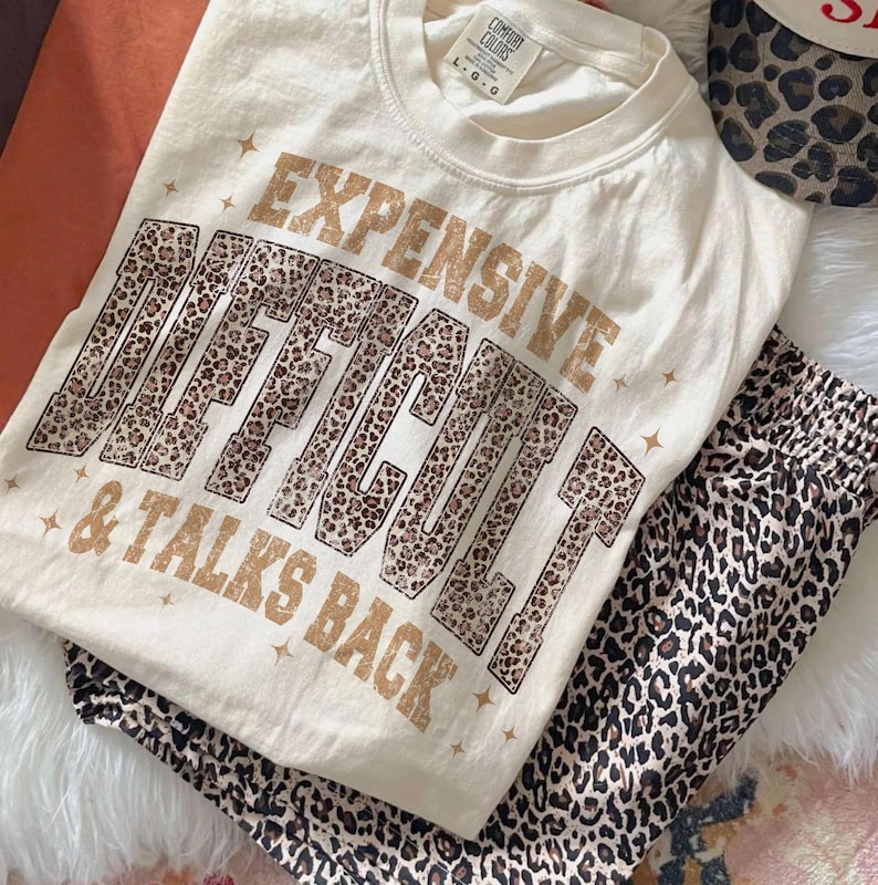May include: Cream-coloured t-shirt with the text "EXPENSIVE, DIFFICULT, & TALKS BACK" in brown and leopard print. The shirt is paired with leopard print trousers. The Comfort Colors brand tag is visible. The outfit is laid out on a white fur-like surface.