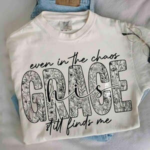 May include: Cream-colored t-shirt with the words "even in the chaos GRACE his still finds me" in black script. The word "GRACE" is filled with a floral pattern. The shirt is folded on top of blue jeans.
