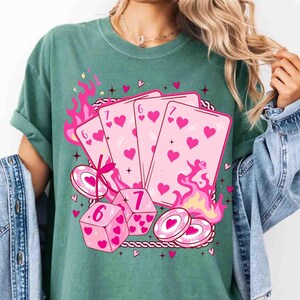 May include: Green t-shirt featuring a pink graphic of playing cards, dice, and poker chips. The cards show hearts and the numbers 6 and 7. Pink flames and hearts surround the design, creating a playful aesthetic.