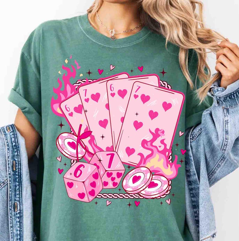 May include: Green t-shirt featuring a pink graphic of playing cards, dice, and poker chips. The cards show hearts and the numbers 6 and 7. Pink flames and hearts surround the design, creating a playful aesthetic.