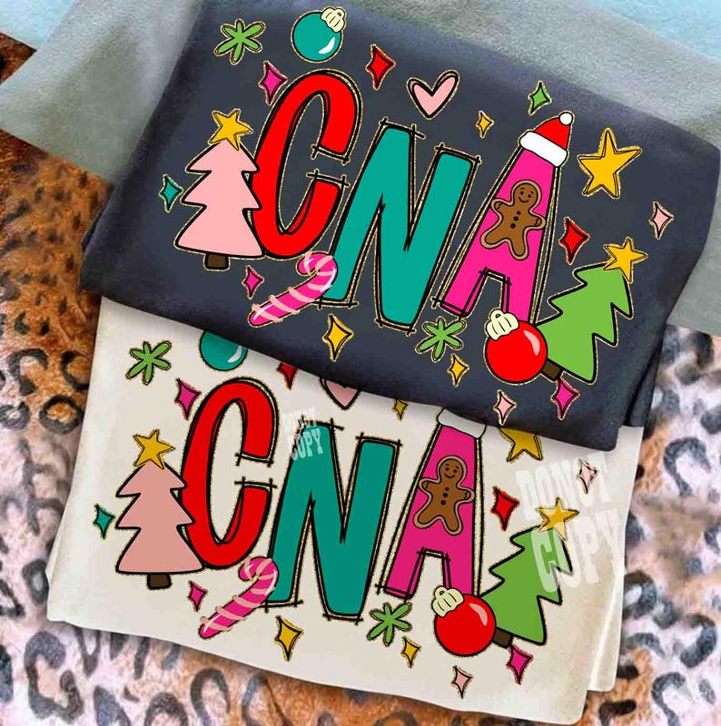May include: Two t-shirts, one dark gray and one off-white, with the word "CNA" in large, colorful letters. The designs include Christmas-themed graphics like trees, ornaments, and gingerbread men. The shirts are folded and stacked.