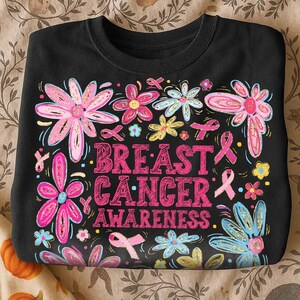May include: Black crewneck sweatshirt with a floral design and the words "Breast Cancer Awareness" in pink. The flowers are pink, blue, and yellow with gold accents, and pink ribbons are scattered throughout.