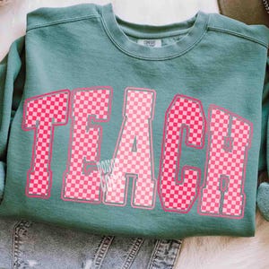 K&ouml;nnte beinhalten: Ein teal-farbenes Sweatshirt mit dem Wort "TEACH" in gro&szlig;en, rosa karierten Buchstaben. Das Sweatshirt hat einen Rundhalsausschnitt und lange &Auml;rmel. Der Text "DON'T COMPROMISE" ist unter dem Buchstaben "A" aufgedruckt.