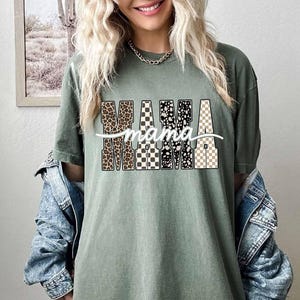 May include: Sage green t-shirt with the word "MAMA" in a bold, patterned design. The letters feature leopard print, black and white checkerboard, and a neutral checkerboard pattern. A cursive "mama" is written across the letters.