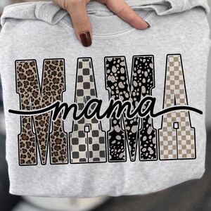 May include: A light gray sweatshirt featuring the word "MAMA" in bold, patterned letters. The letters have a leopard print, checkered, and spotted design. The word "mama" is written in a cursive font underneath the word "MAMA".