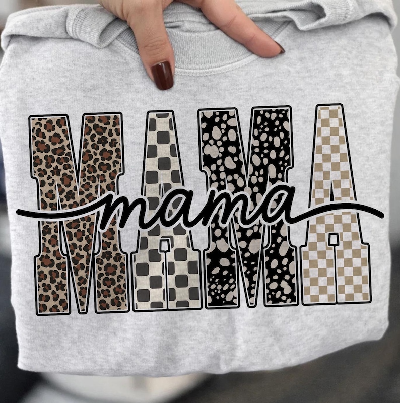 May include: A light gray sweatshirt featuring the word "MAMA" in bold, patterned letters. The letters have a leopard print, checkered, and spotted design. The word "mama" is written in a cursive font underneath the word "MAMA".