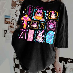 May include: Black t-shirt with a colorful Easter-themed graphic. The design includes a cross, Easter eggs, bunnies, bows, and the words "Happy" and "Easter". The graphic is framed with neon colors.