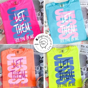 May include: Four colorful t-shirts: pink, blue, orange, and neon yellow. Each shirt features the text "LET THEM SEE YOU IN ME" in a contrasting color. The shirts are displayed with disco balls and a magazine.