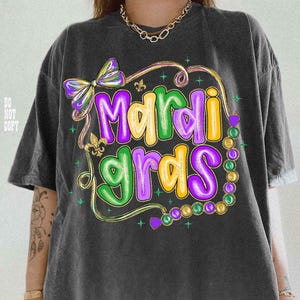 May include: Dark gray t-shirt with a colorful "Mardi Gras" graphic. The design features purple, green, and yellow text, a bow, and a strand of beads. The shirt has a relaxed fit.
