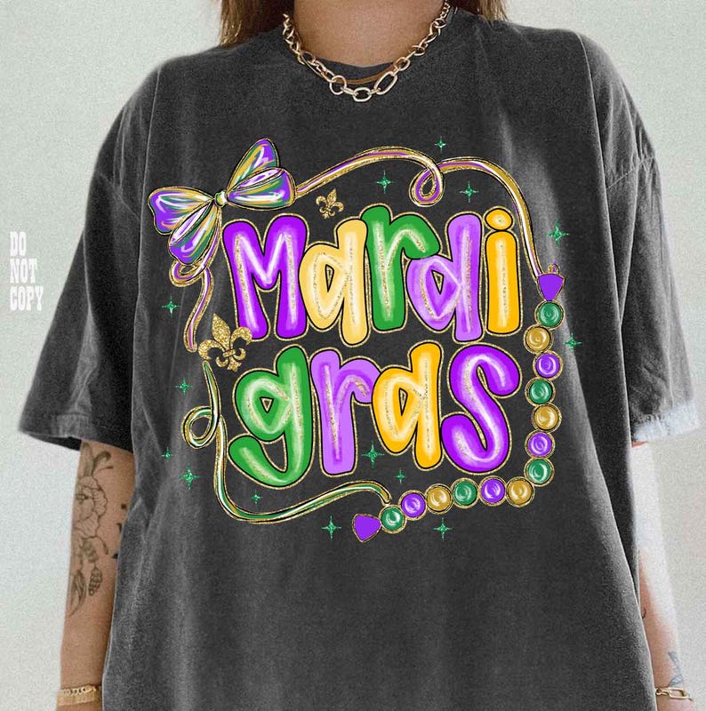 May include: Dark gray t-shirt with a colorful "Mardi Gras" graphic. The design features purple, green, and yellow text, a bow, and a strand of beads. The shirt has a relaxed fit.