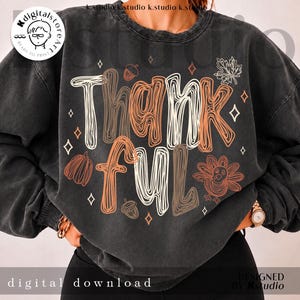 K&ouml;nnte beinhalten: Ein anthrazitfarbenes Sweatshirt mit Rundhalsausschnitt und dem Wort Thankful in einer stilisierten, mehrzeiligen Schriftart. Das Design enth&auml;lt orangefarbene und wei&szlig;e grafische Illustrationen eines Truthahns, eines K&uuml;rbisses, Eicheln, Bl&auml;ttern und kleinen Rauten.