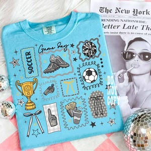 May include: A light blue t-shirt with a soccer theme. The shirt features graphics of soccer cleats, a soccer ball, a trophy, a whistle, and the words "Game Day" and "Soccer". The shirt also has star and bow accents.