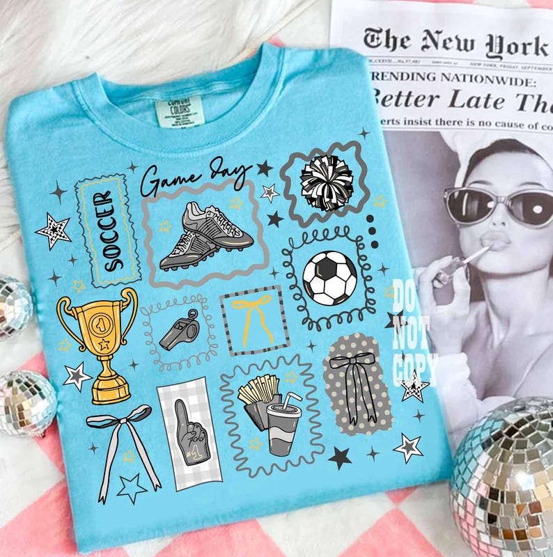 May include: A light blue t-shirt with a soccer theme. The shirt features graphics of soccer cleats, a soccer ball, a trophy, a whistle, and the words "Game Day" and "Soccer". The shirt also has star and bow accents.