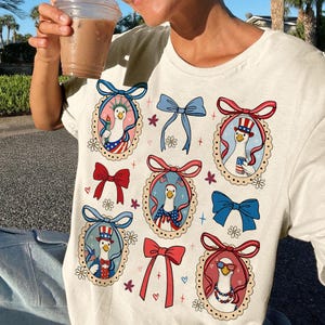 Op de afbeelding: Cr&egrave;mekleurig T-shirt met patriottische ganzenillustraties in ovale frames met strikken. De ganzen zijn gekleed in Amerikaanse vlag-thema kleding, houden een fakkel vast en dragen een zonnebril. Het shirt heeft een casual, feestelijk ontwerp.