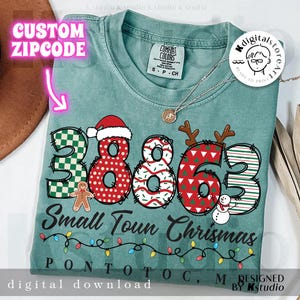 Op de afbeelding: Een teal Comfort Colors t-shirt met een kerstthema ontwerp. Het ontwerp bevat de nummers "38863" met feestelijke patronen, de woorden "Small Town Chrismas" en de plaatsnaam "Pontotoc, M". Een digitale download.