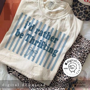 May include: Cream-colored t-shirt with the text "I'd rather be Thrifting" in blue, accented by blue vertical stripes and a bow graphic. Leopard print pants and a leopard print hat are also visible. The image also includes the text "digital download".