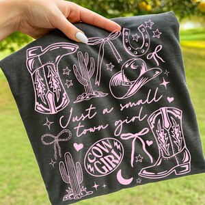 May include: Dark gray t-shirt with a pink graphic design featuring cowboy boots, a horseshoe, a cowboy hat, cacti, bows, stars, and the words "Just a small town girl" and "Cow Girl".