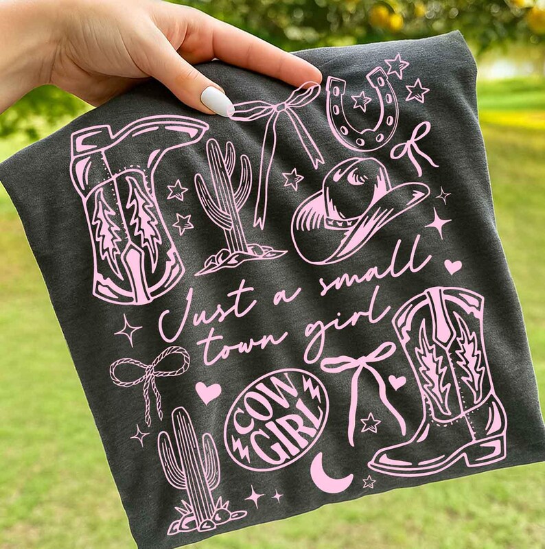 May include: Dark gray t-shirt with a pink graphic design featuring cowboy boots, a horseshoe, a cowboy hat, cacti, bows, stars, and the words "Just a small town girl" and "Cow Girl".