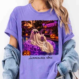 May include: A purple t-shirt with a graphic of a ghost wearing sunglasses, relaxing in a pool on a purple inflatable ring. The image includes jack-o'-lanterns and the text "Summerween vibes."
