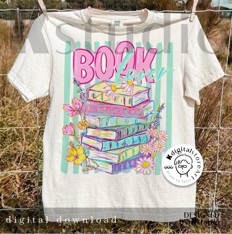 May include: Cream-colored t-shirt with a colorful graphic of stacked books, flowers, and the words "BOOK Lover". The design features pink, blue, yellow, and green hues. The shirt is hanging on a clothesline.