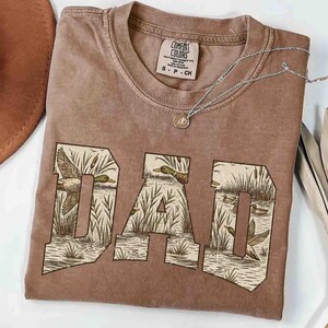 May include: A brown Comfort Colors t-shirt with the word "DAD" in a block letter design. The letters are filled with a duck and cattail scene. A silver necklace with a small pendant rests on the shirt.