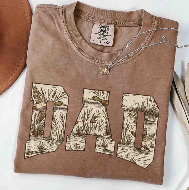 May include: A brown Comfort Colors t-shirt with the word "DAD" in a block letter design. The letters are filled with a duck and cattail scene. A silver necklace with a small pendant rests on the shirt.