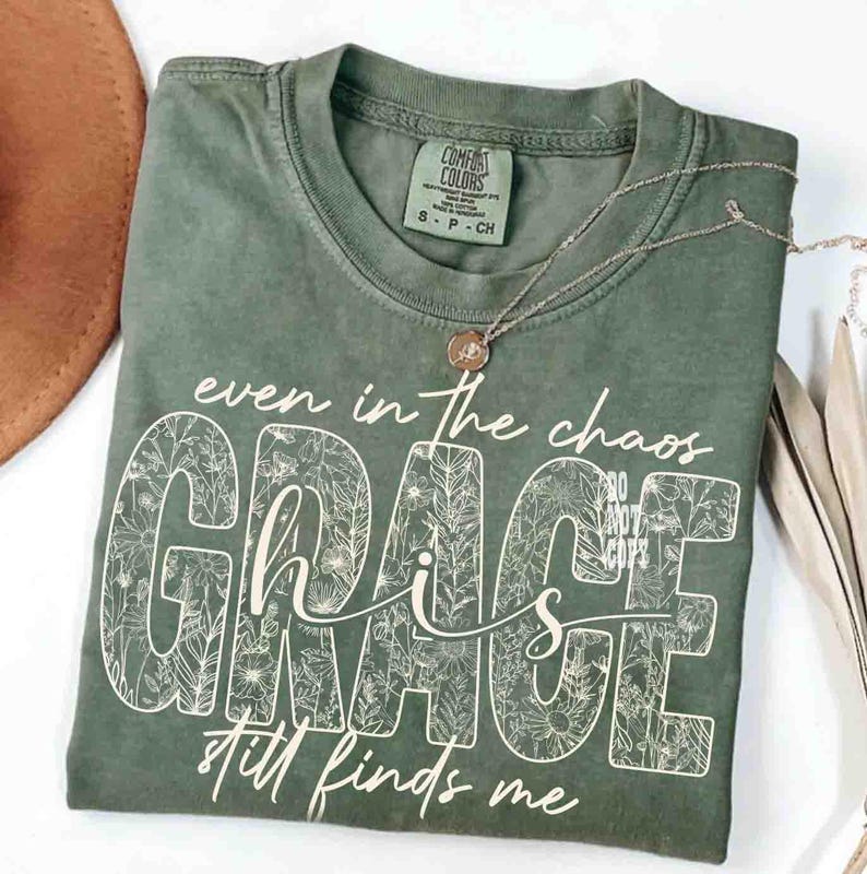 May include: An olive green t-shirt with the words "even in the chaos GRACE still finds me" in white script and floral design. A gold necklace with a pendant rests on the shirt. The shirt is folded and next to a brown hat.