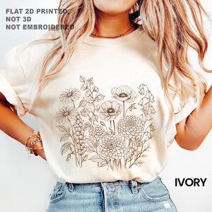 May include: Ivory-colored t-shirt featuring a detailed floral line drawing. The design includes daisies, poppies, and other flowers. The text "FLAT 2D PRINTED, NOT 3D, NOT EMBROIDERED" is printed at the top, and "IVORY" is printed at the bottom.