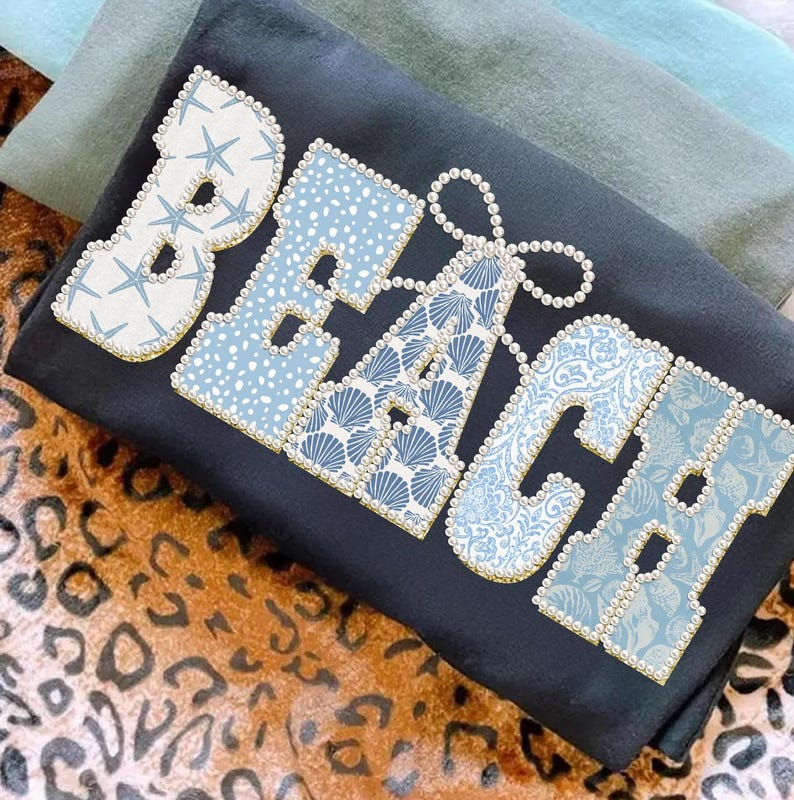 May include: A black t-shirt featuring the word "BEACH" in large, decorative letters. The letters are filled with patterns like starfish, polka dots, and seashells, and are outlined with pearls. The shirt is folded, with other fabric visible.
